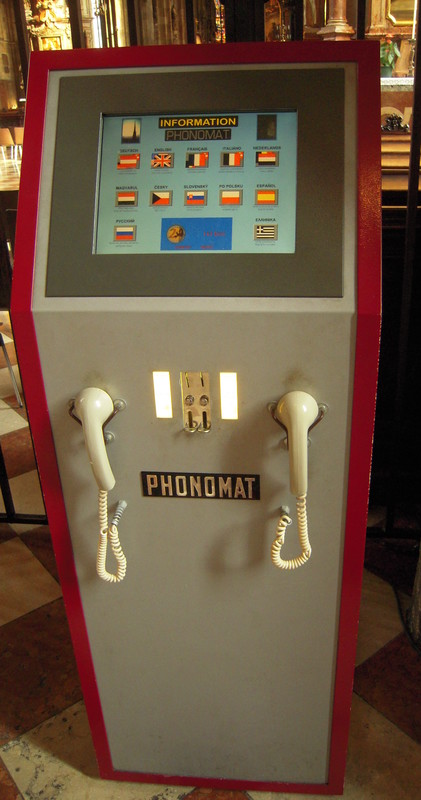 A tall, floor–standing box labeled PHONOMAT, with two telephone receivers
	(lacking mouthpiece) hanging side–by–side on the front, slots for accepting Euro coins,
	and a touch–screen on the angled top offering German, English, French, Italian, Dutch, Hungarian, Czech, Slovak,
	Polish, Spanish, Russian, and Greek languages.